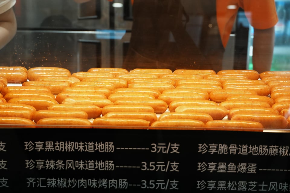 Rows of grilled sausages behind a priced display counter.