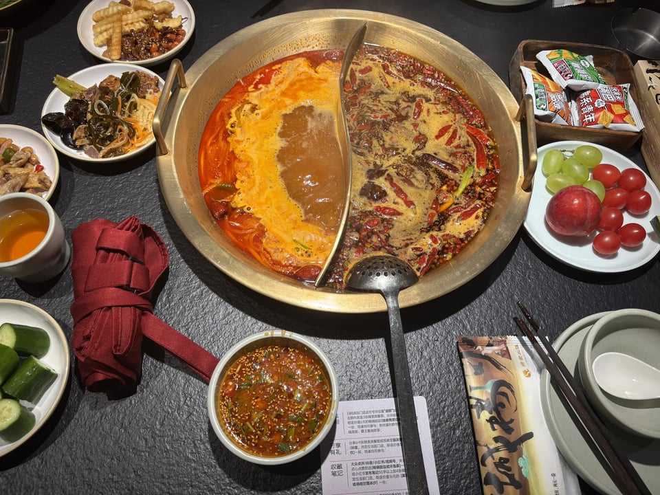 A divided pot of spicy hot pot surrounded by side dishes and fruit.
