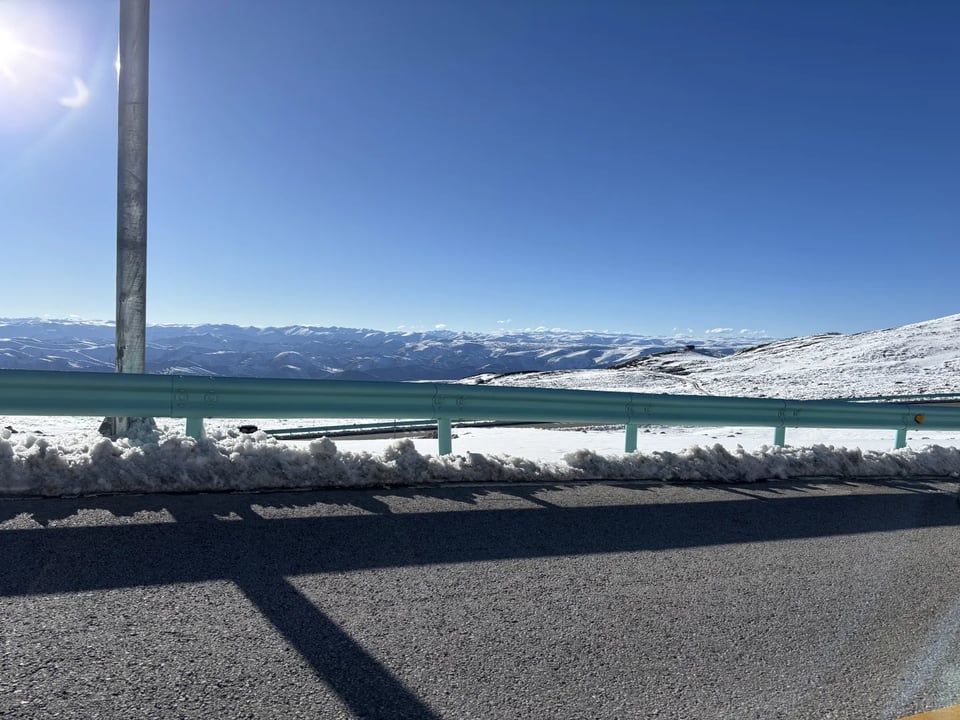 A snowy plateau and distant ranges seen past a mountain road guardrail.