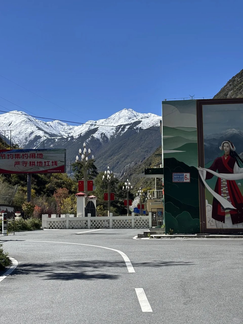Snow-capped peaks behind a mountain road and a mural of a Tibetan woman.