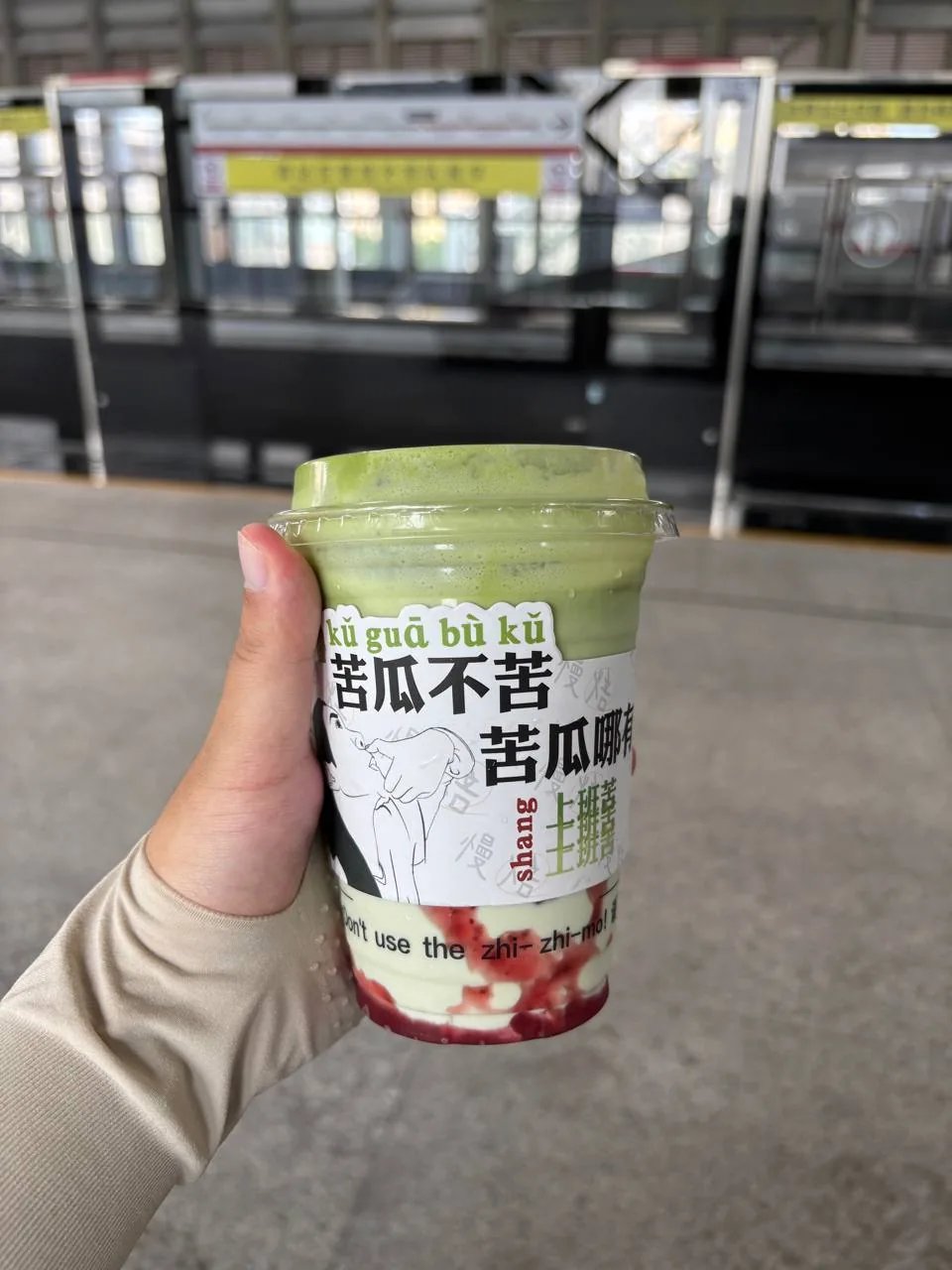 A hand holds a green drink in a printed cup on a metro platform.
