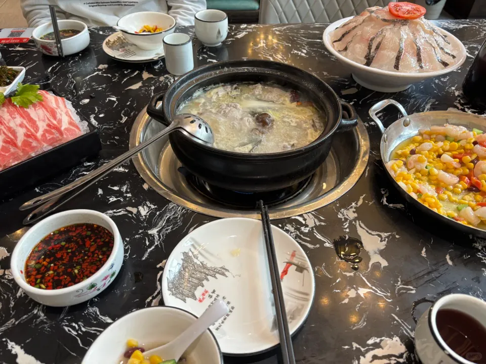 A hotpot table with fish soup, sliced beef, shrimp with corn, and dipping sauces.