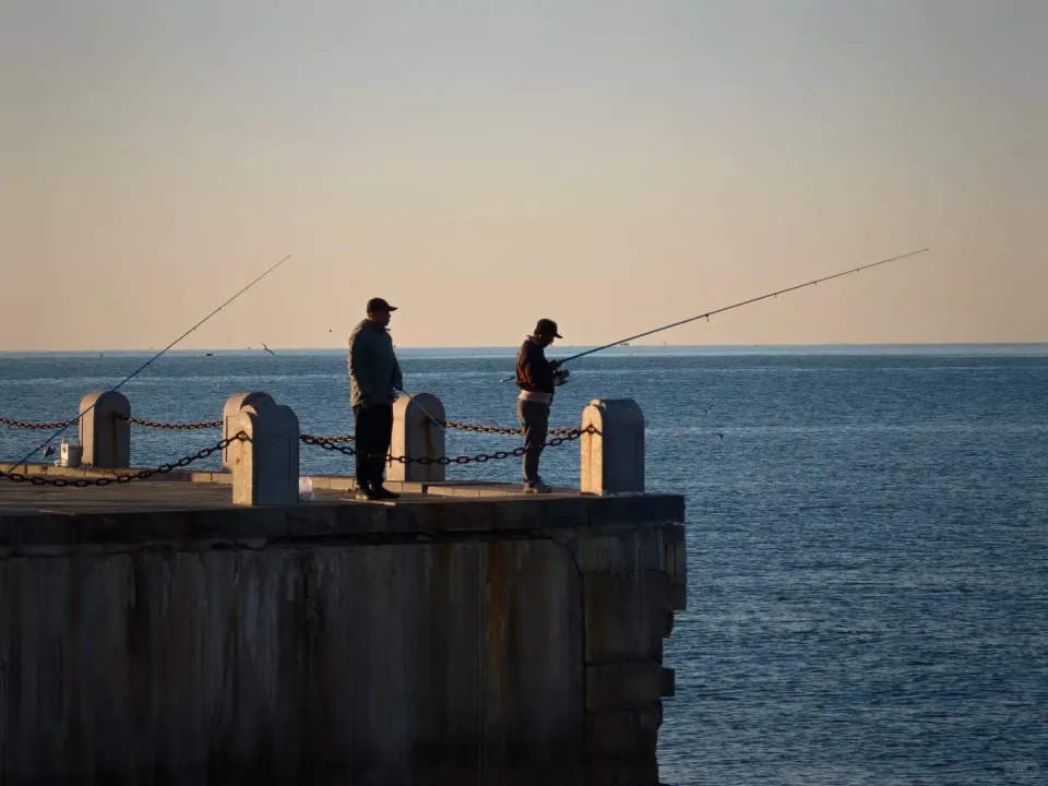 Two people fishing from a seaside platform in the morning light.