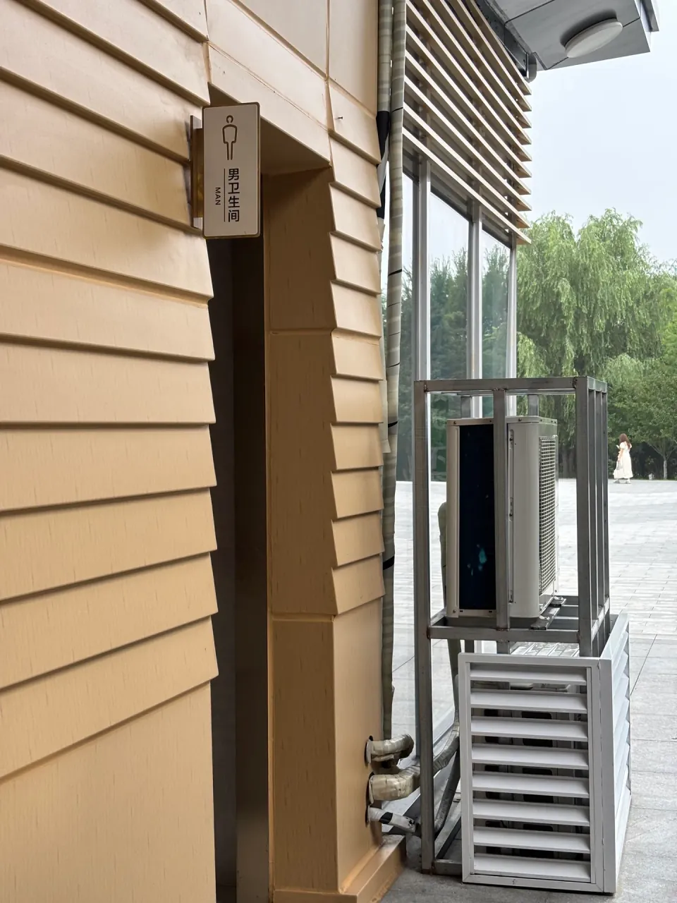 The entrance to a men's restroom beside ventilation equipment and trees.