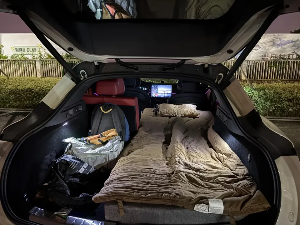 An open car hatch revealing a mattress, bedding, and bags arranged for sleeping.