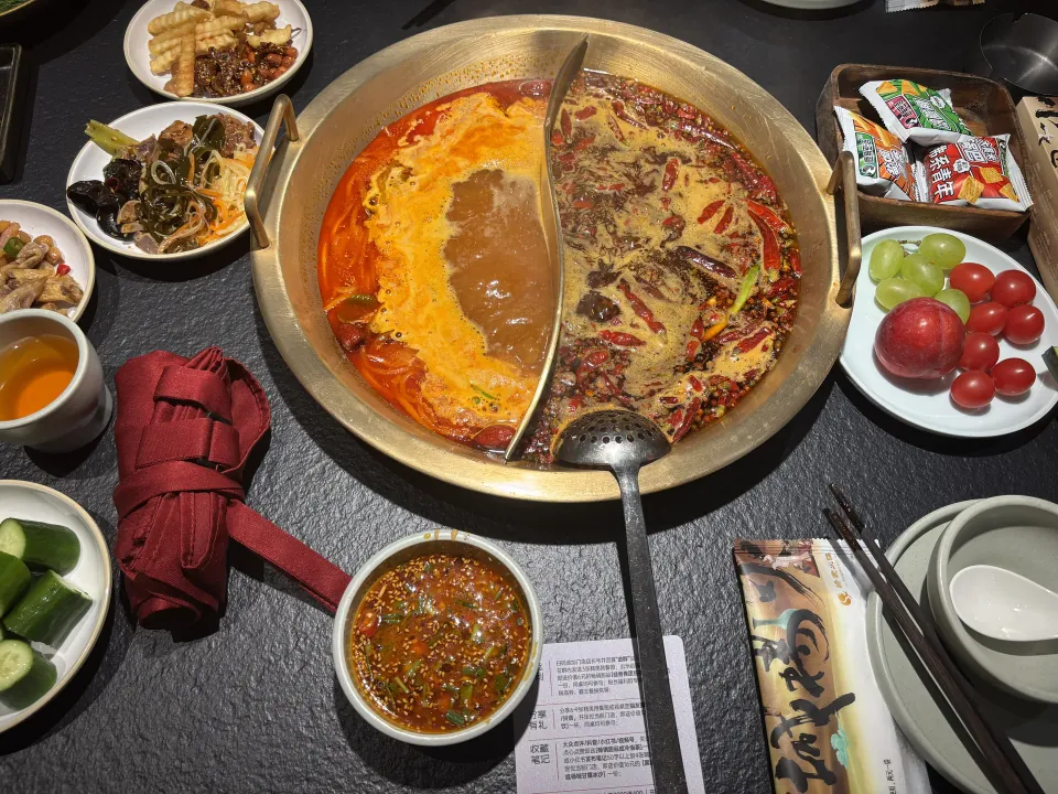 A divided pot of spicy hot pot surrounded by side dishes and fruit.