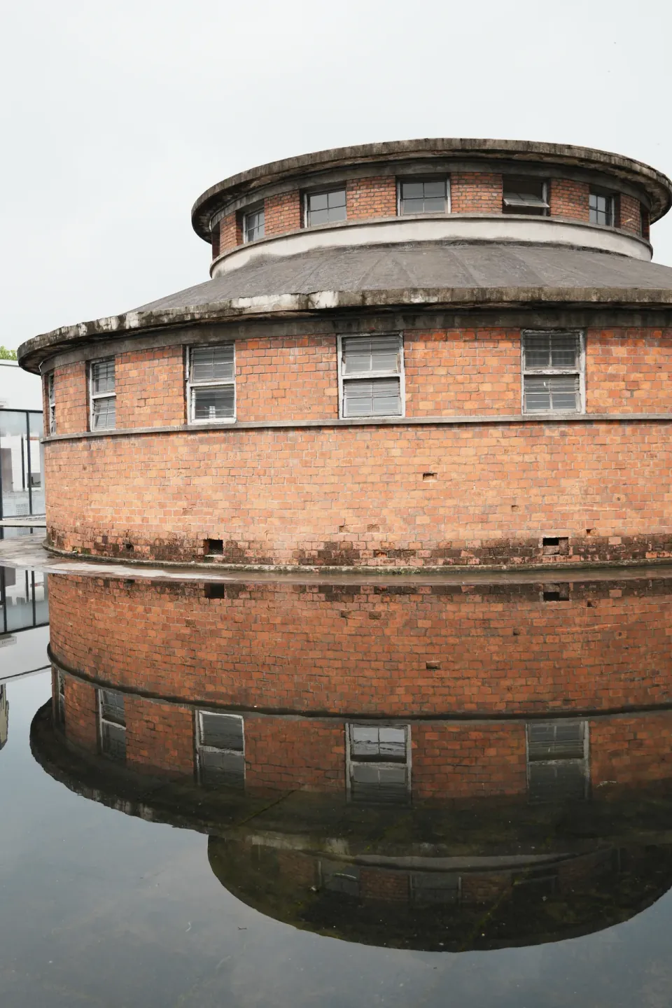 A round red-brick industrial building reflected in a shallow pool.
