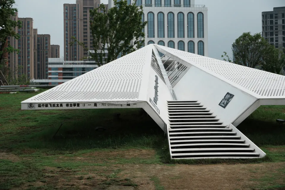 A white geometric pavilion on a lawn with apartment buildings behind it.
