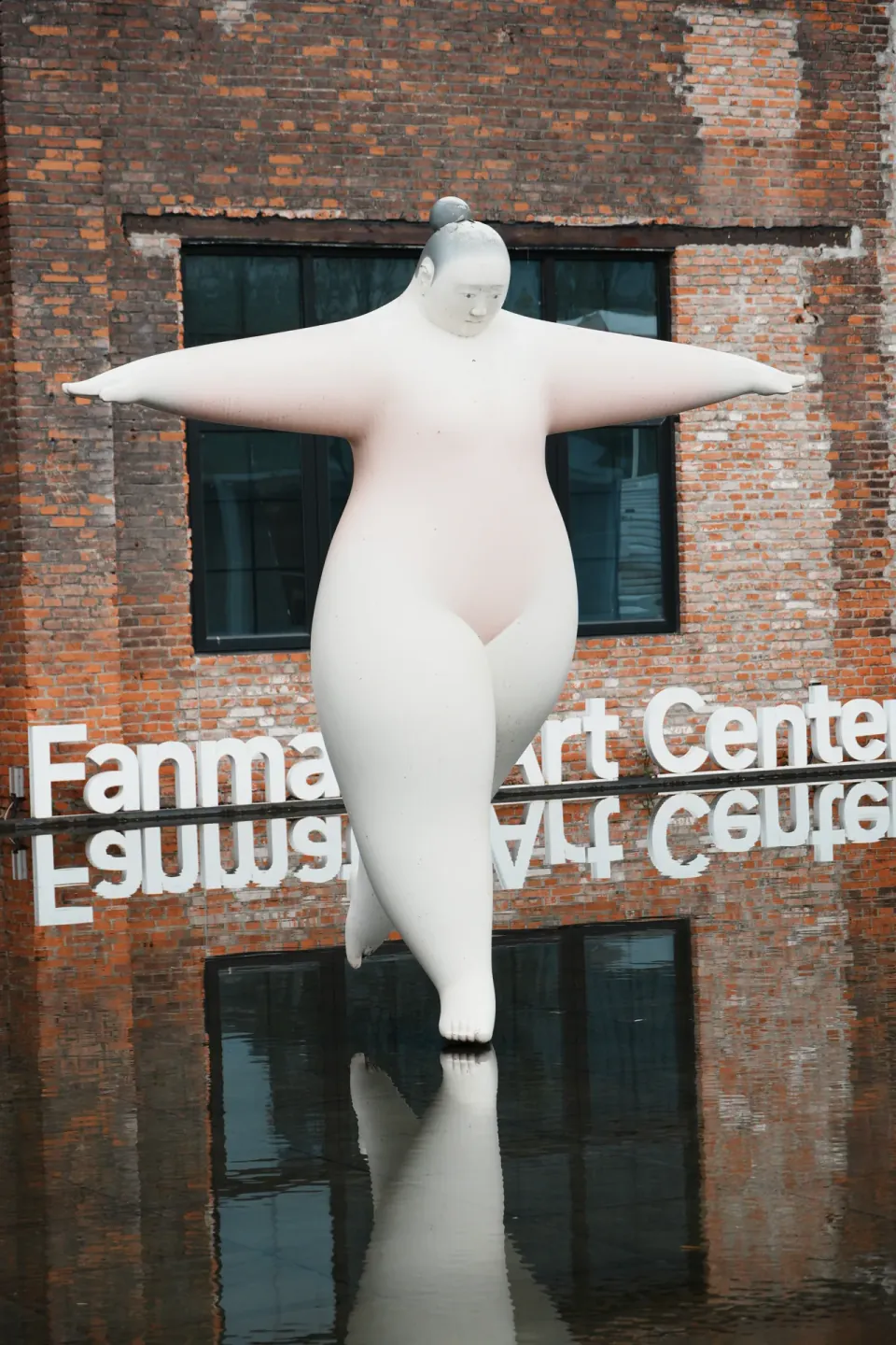 A large white figure sculpture reflected in water outside a brick art center.