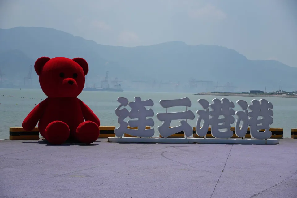 Red teddy bear beside a Lianyungang Harbor sign overlooking the sea