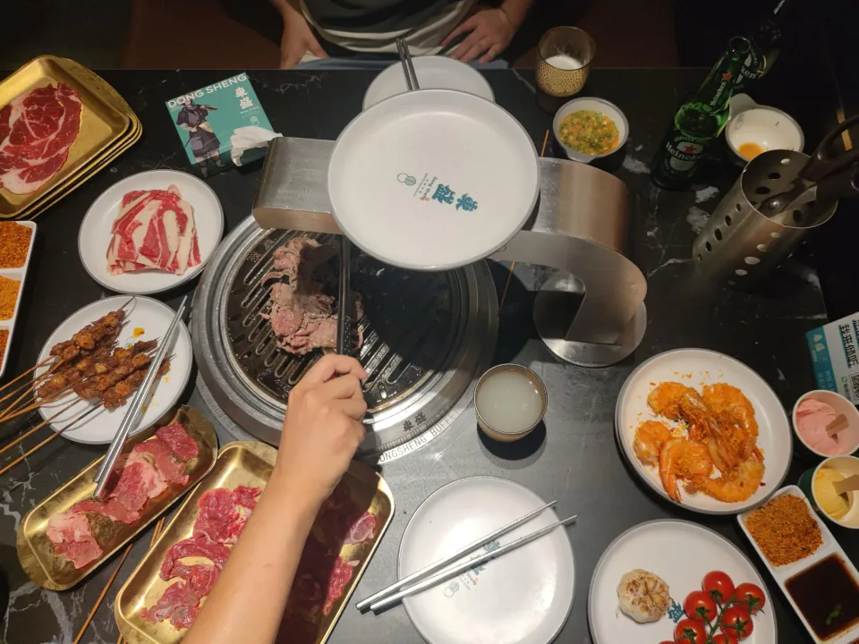 Diners grill meat at a table covered with plates of barbecue ingredients and side dishes.