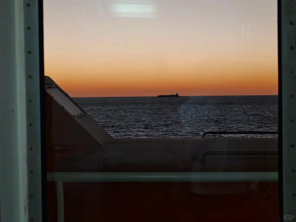 A distant ship at dusk, seen past the ferry's railing and window frame.