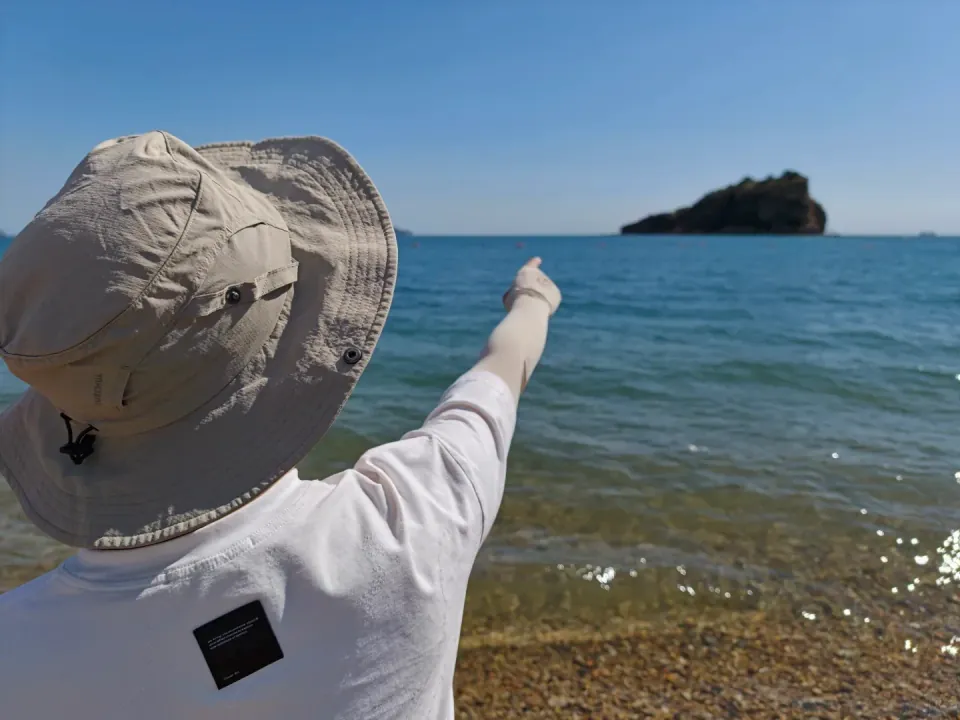A person in a sun hat pointing toward an island from the beach.