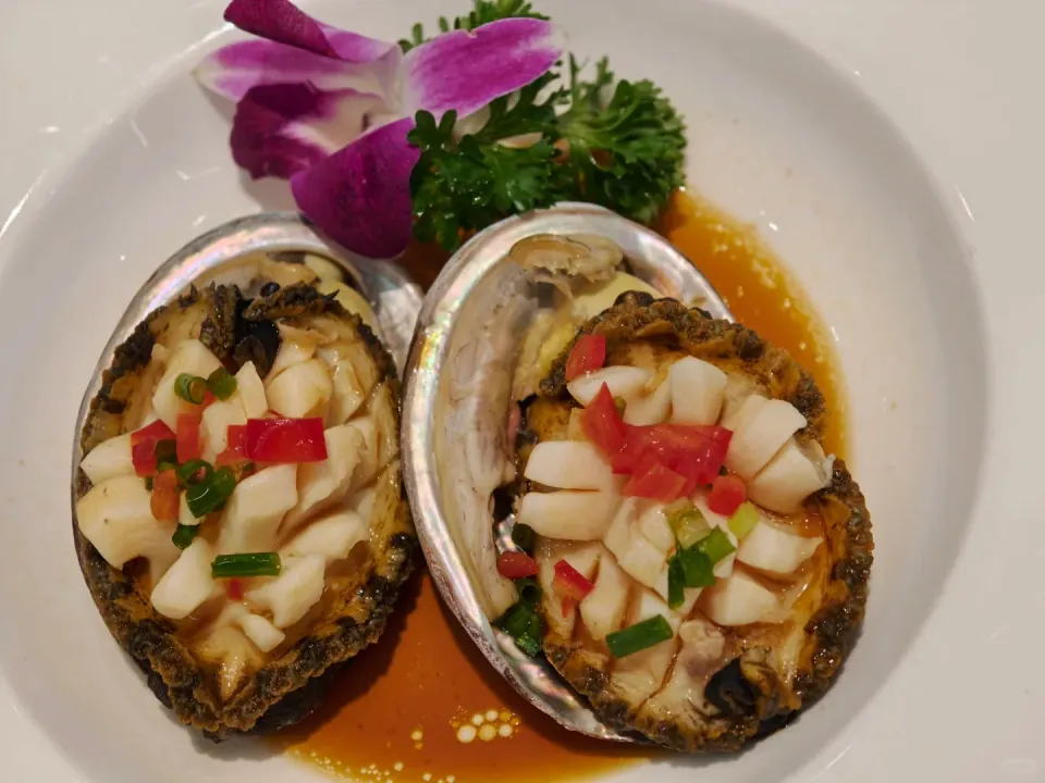 Two abalone in their shells with chopped garnishes and sauce.