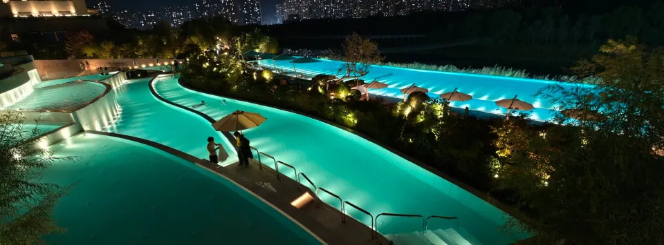 A wide night view of illuminated pools and distant city buildings.